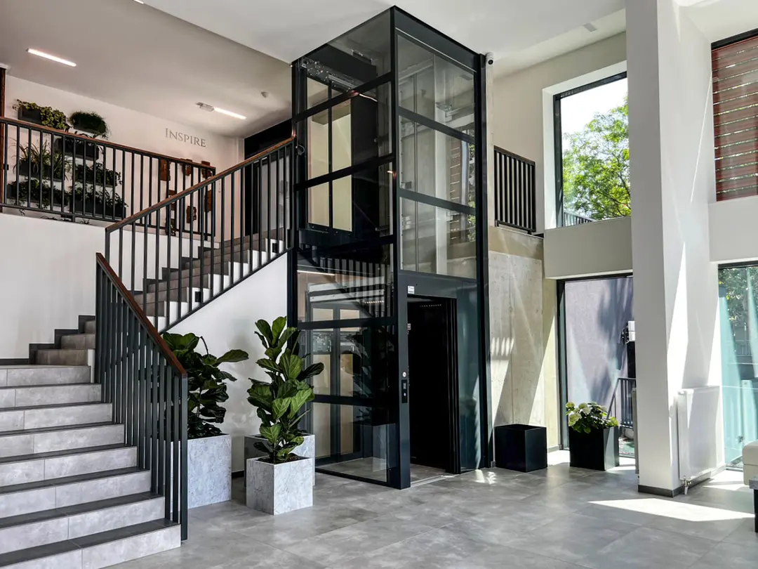 Cibes C1 Pure cabin lift with a black framed glass shaft installed in a bright lobby stairwell, ground-floor landing open.