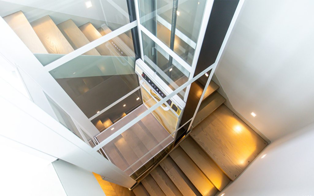 Cibes home lift with glass shaft fitted in a stairwell, viewed from above with warm step lighting.