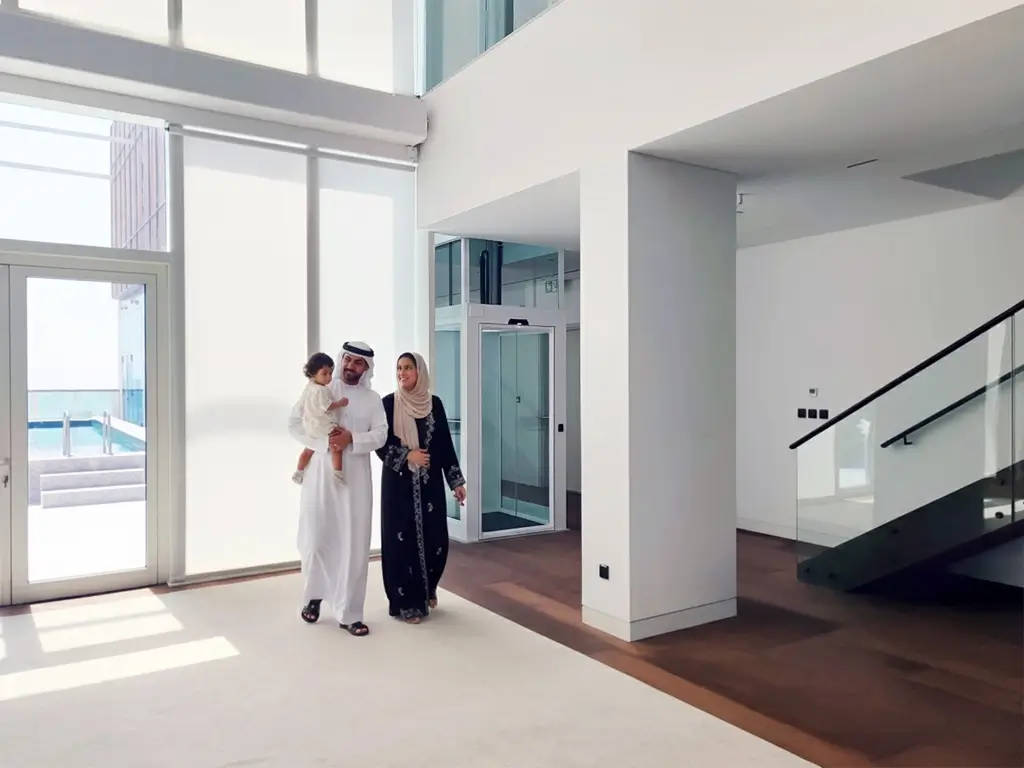 Emirati couple with a child walking in a bright Dubai villa; a Cibes home lift with glass panels stands beside the staircase.