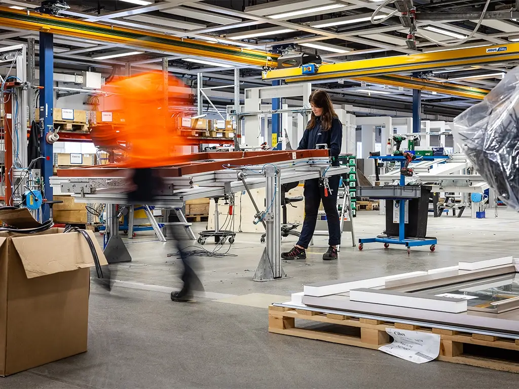 Technician assembling a home-elevator frame on a factory line; another worker in an orange jacket passes by; pallets and overhead cranes visible.