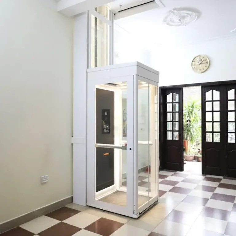 The white, compact Ascenda Minivator home lift with glass panels, installed in the corner of a room with a checkered tile floor.